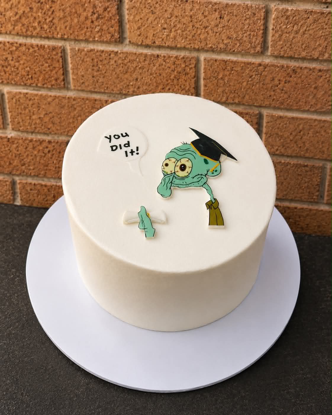White smooth buttercream graduation cake with Squidward in graduation cap edible print and You Did It speech bubble